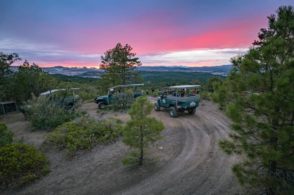 Off-road vehicles parked on a winding dirt path with a colorful sunset and mountains in the background.