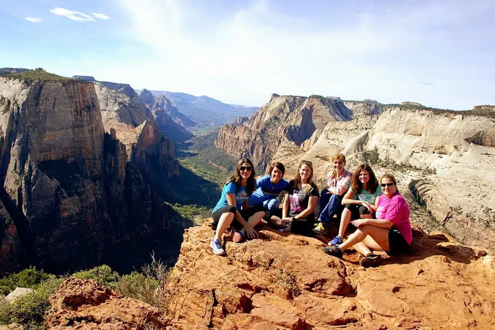 A group of six women sits on a rock ledge overlooking a stunning canyon landscape.