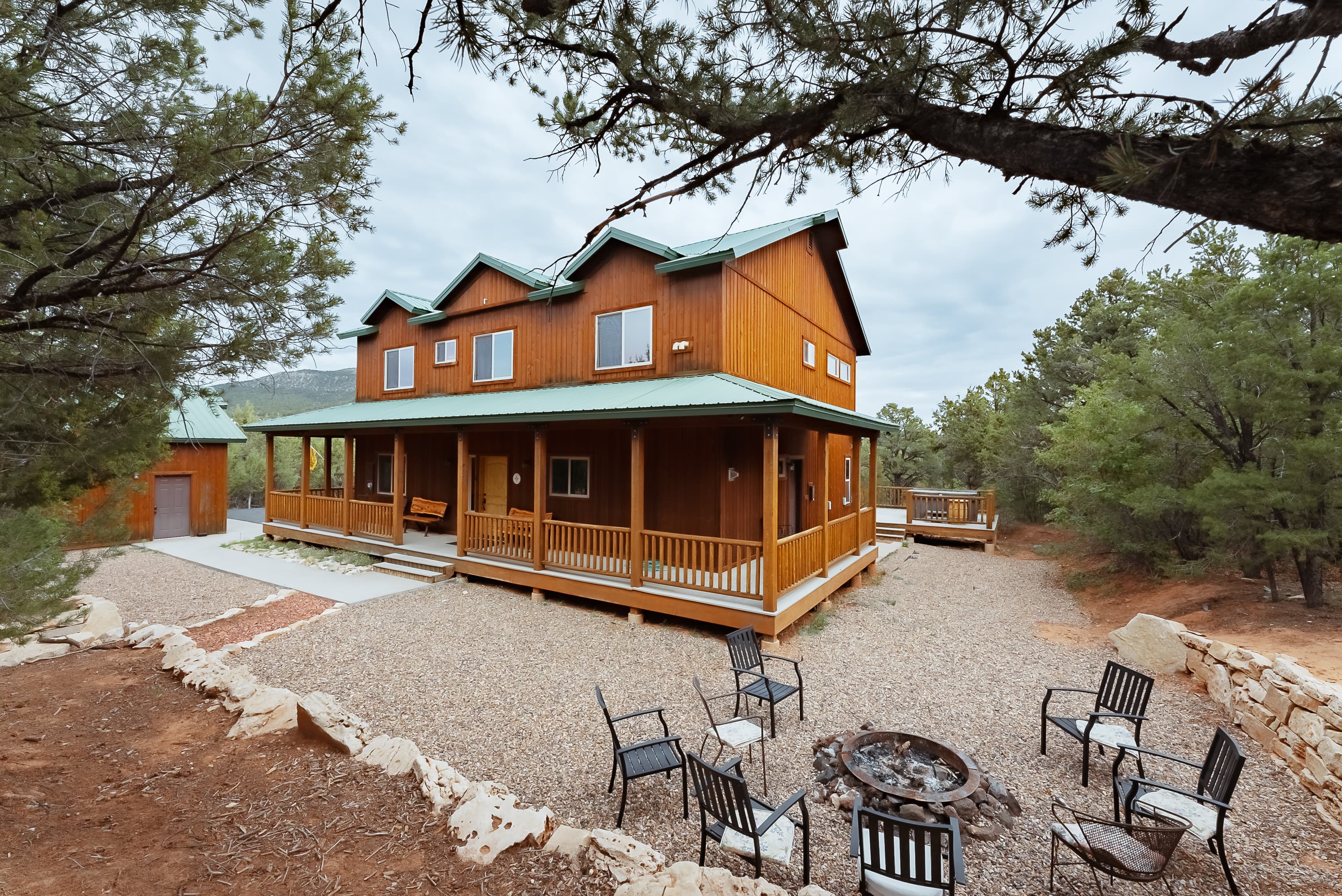 A wooden cabin with a green roof, surrounded by trees, features a patio and a fire pit with chairs in a gravel area.