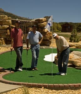 Three men are standing on a miniature golf course, with one preparing to putt near the hole.