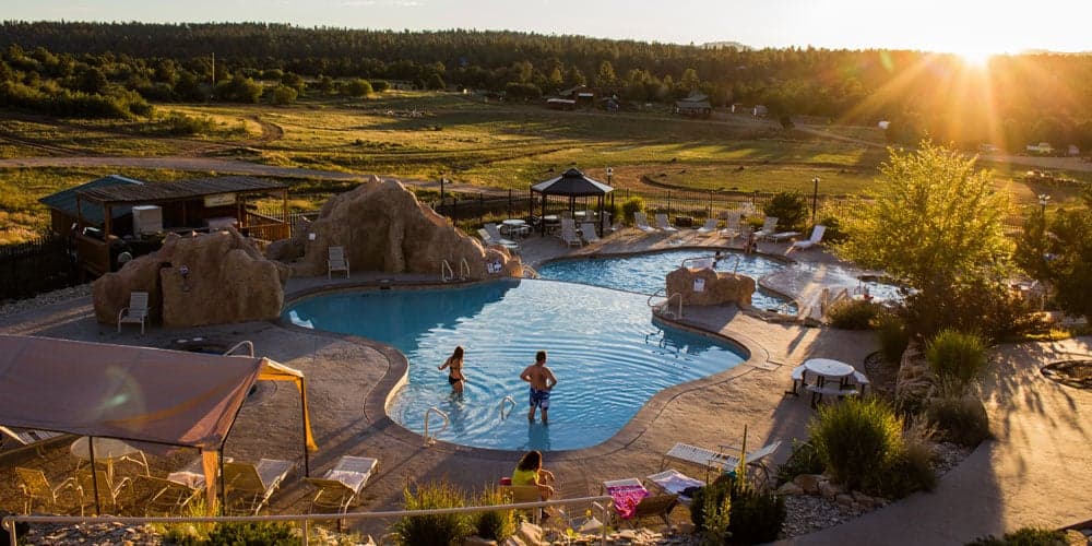 A scenic view of a pool surrounded by rocky formations and lush greenery at sunset, with two people enjoying the water.