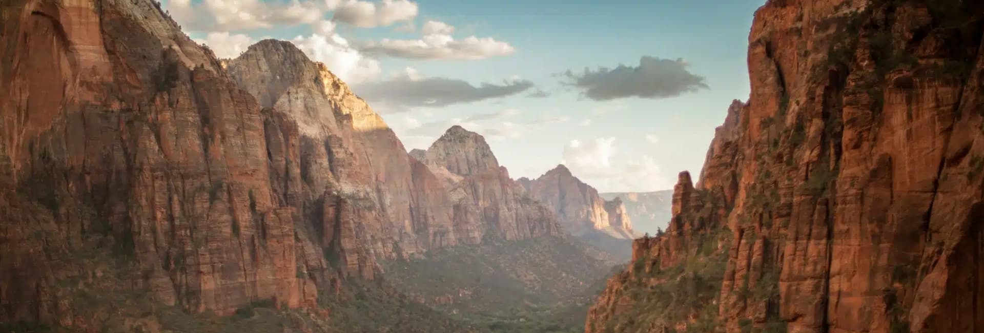 A panoramic view of rugged red cliffs and mountains under a blue sky with scattered clouds.