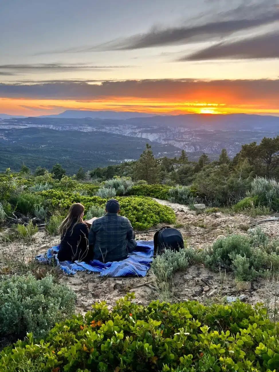 A couple sits on a blanket overlooking a sunset in a lush, mountainous landscape.
