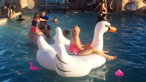a teenage girl sits on an inflatable goose in a swimming pool while younger children and their fathers play chicken in the pool behind her