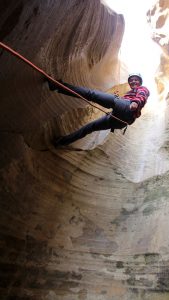 an adventurer smiles down at camera while suspended on a rock wall near zion national park
