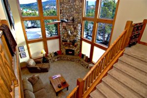 A view from the top of the stairs of a rental home at zion ponderosa ranch look down upon a warm living room with a stone fireplace and a snowy view out of the windows.