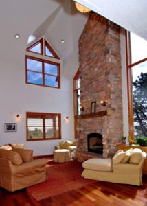 A large living room has vaulted ceilings, floor to ceiling windows, a large stone brick fireplace, and several sofas